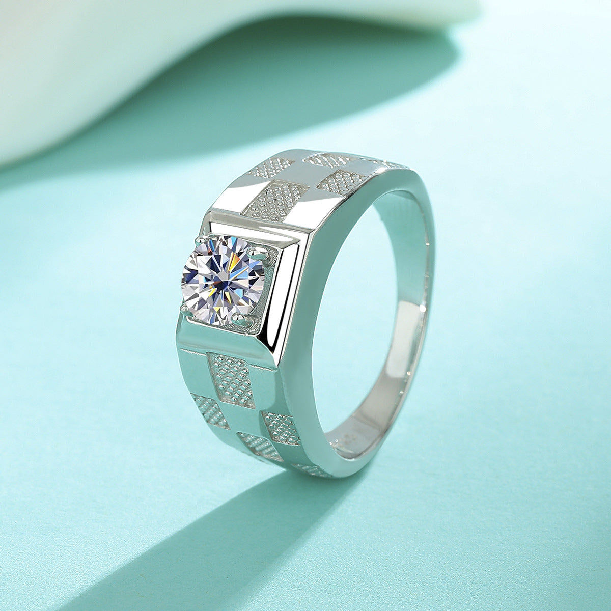 A silver geometric ring with a round-cut clear gemstone, displayed on a light blue surface with a soft white background.