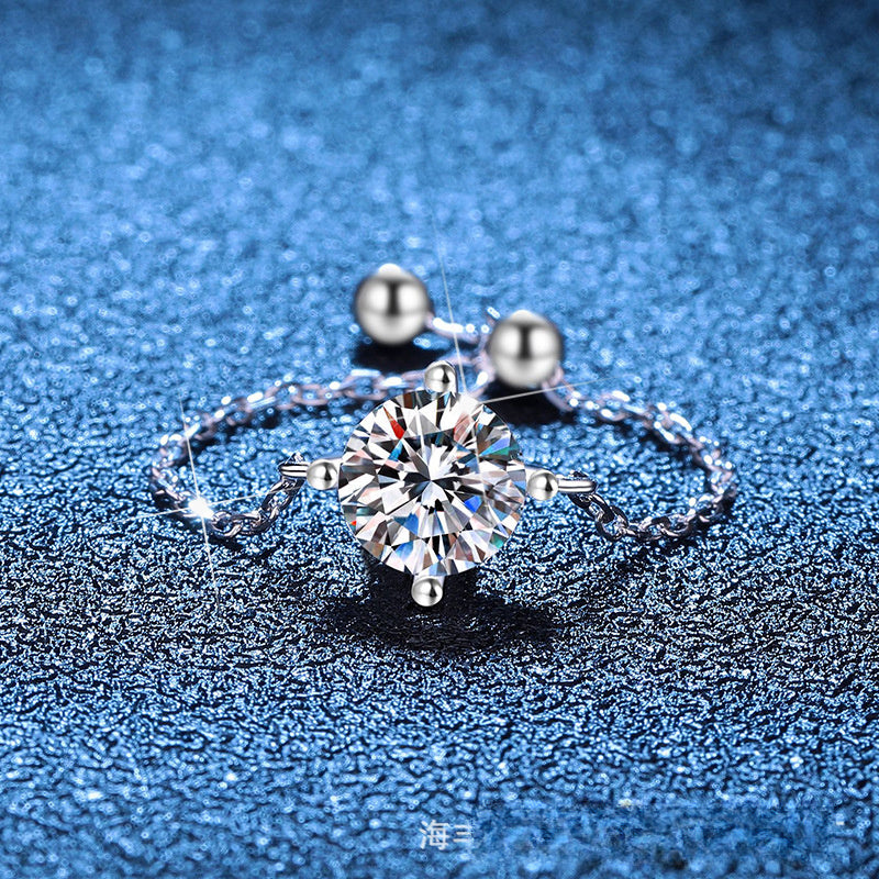 A silver-toned ring with a large round clear gemstone in a prong setting, shown on a textured blue background. The ring has small bead details on the band and a small silver charm attached to the ring, photographed close-up.