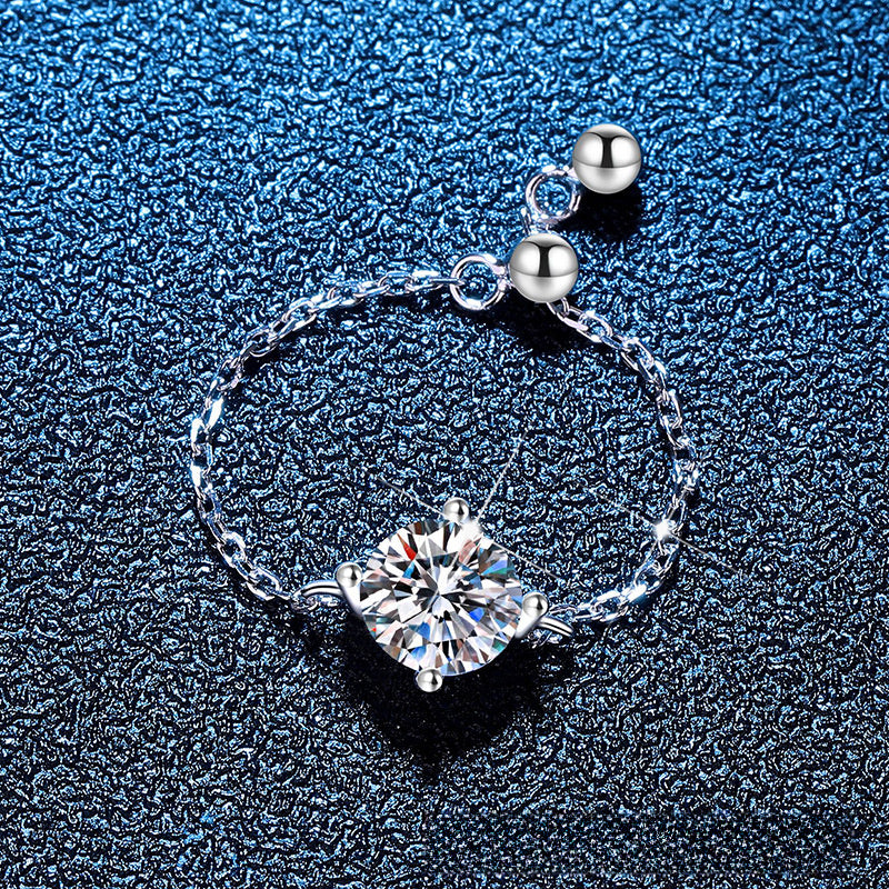 A silver chain bracelet featuring a large clear square-cut gemstone in a prong setting, with two small polished silver beads at the clasp, displayed on a dark textured blue background.