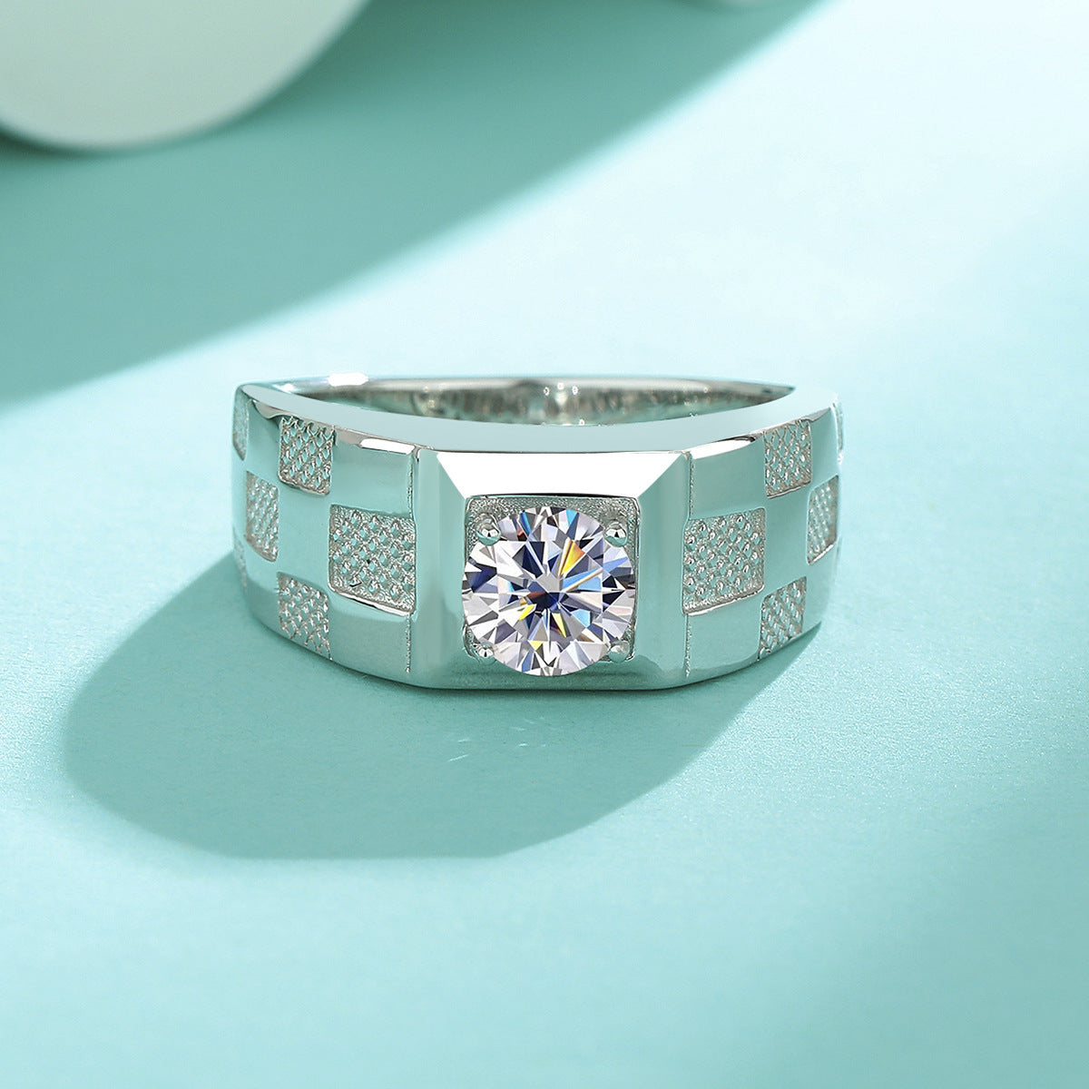 A silver-toned men's ring with a large round clear gemstone and textured geometric side panels, photographed on a light aqua background.