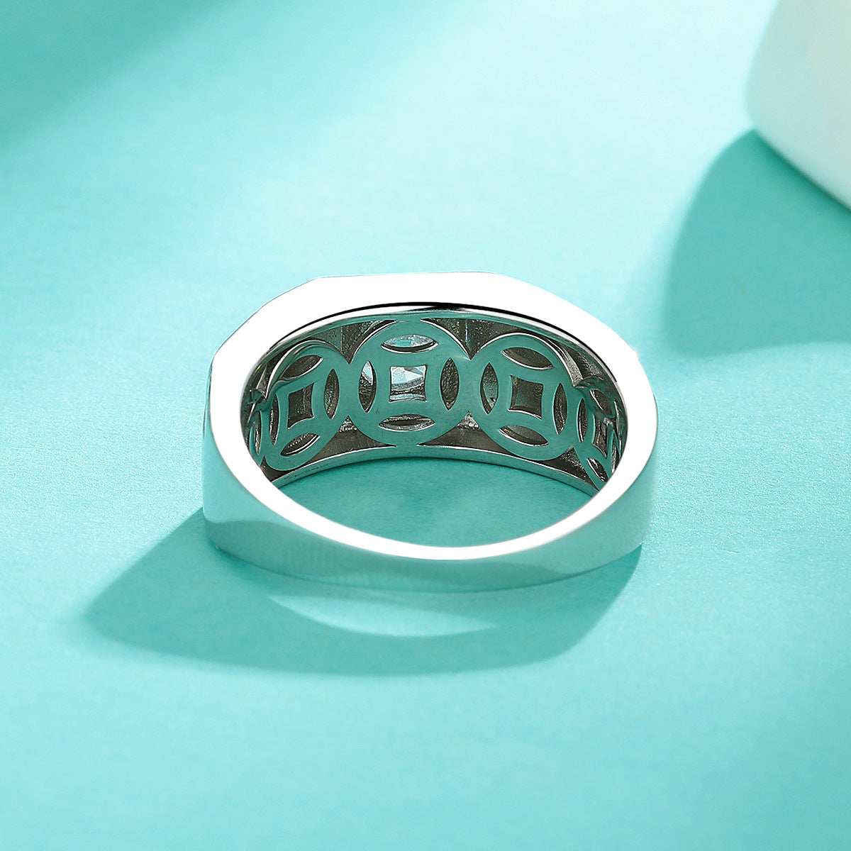A silver-toned ring with a wide band and an openwork circular cutout pattern on the inside, photographed on a smooth aqua background.