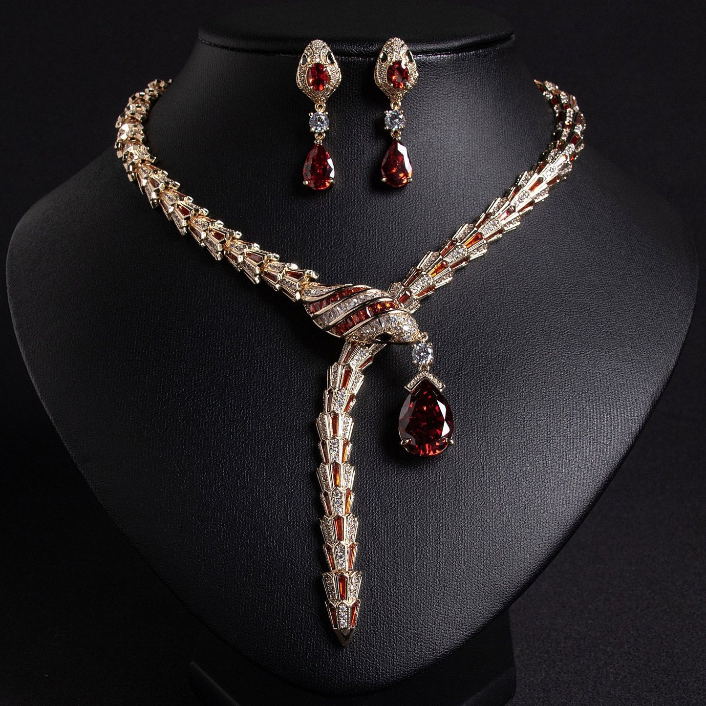 Gold snake necklace and earrings set with red and clear stones, on a black bust.