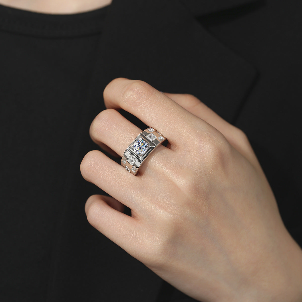 A silver-toned ring with a rectangular bezel-set clear gemstone and a textured band, shown worn on a person's hand against a black shirt background.