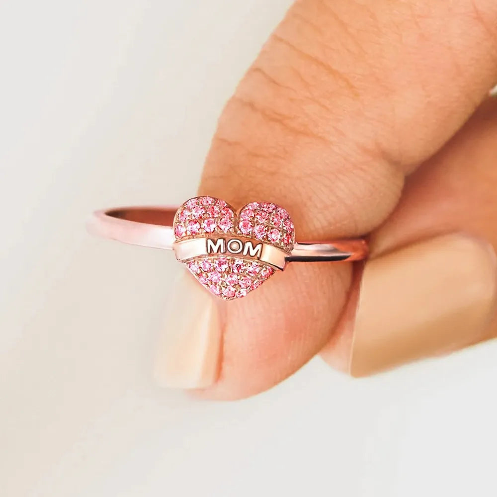 Heart-shaped ring with 'MOM' engraving held between fingers on a light background