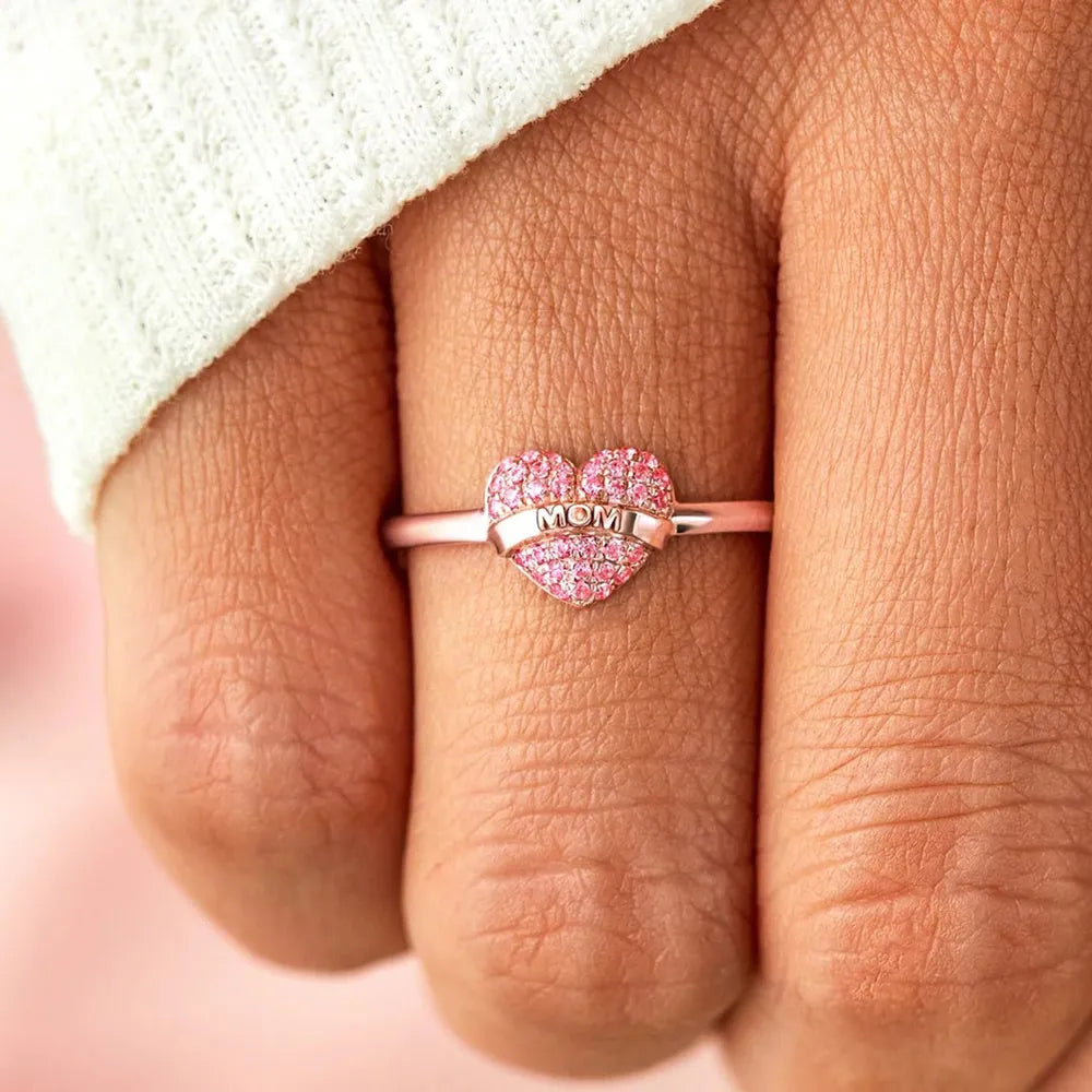 Heart-shaped ring with pink stones and 'MOM' engraving on a finger.