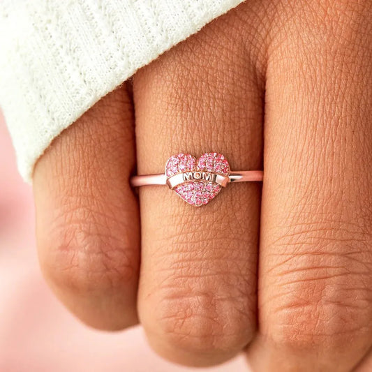 Heart-shaped ring with pink stones and 'MOM' engraving on a finger.