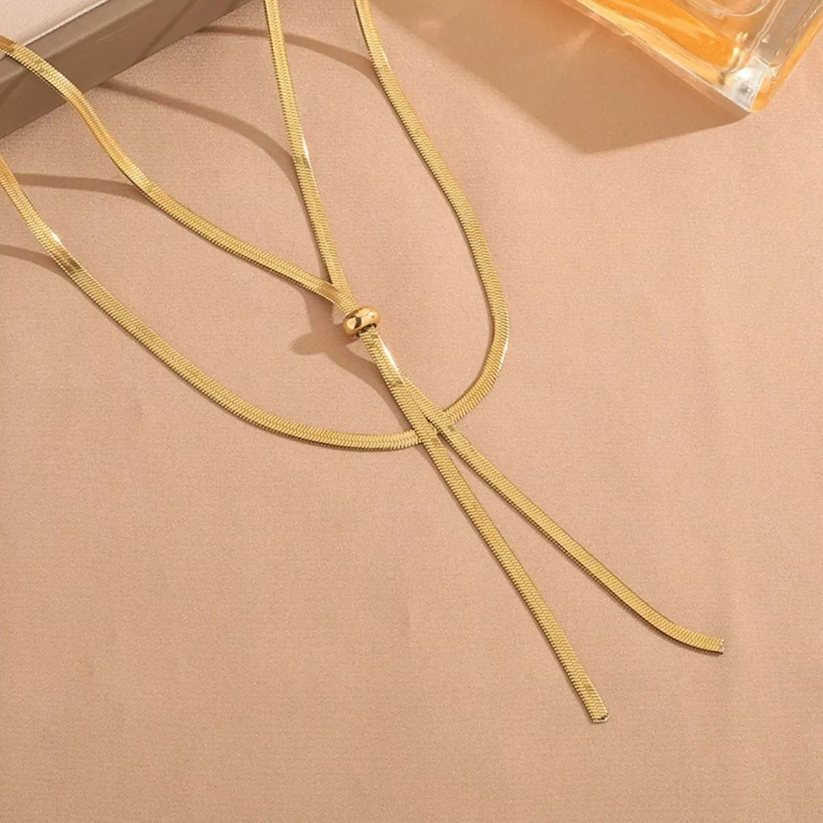 Gold necklace on a beige surface with a perfume bottle in the background