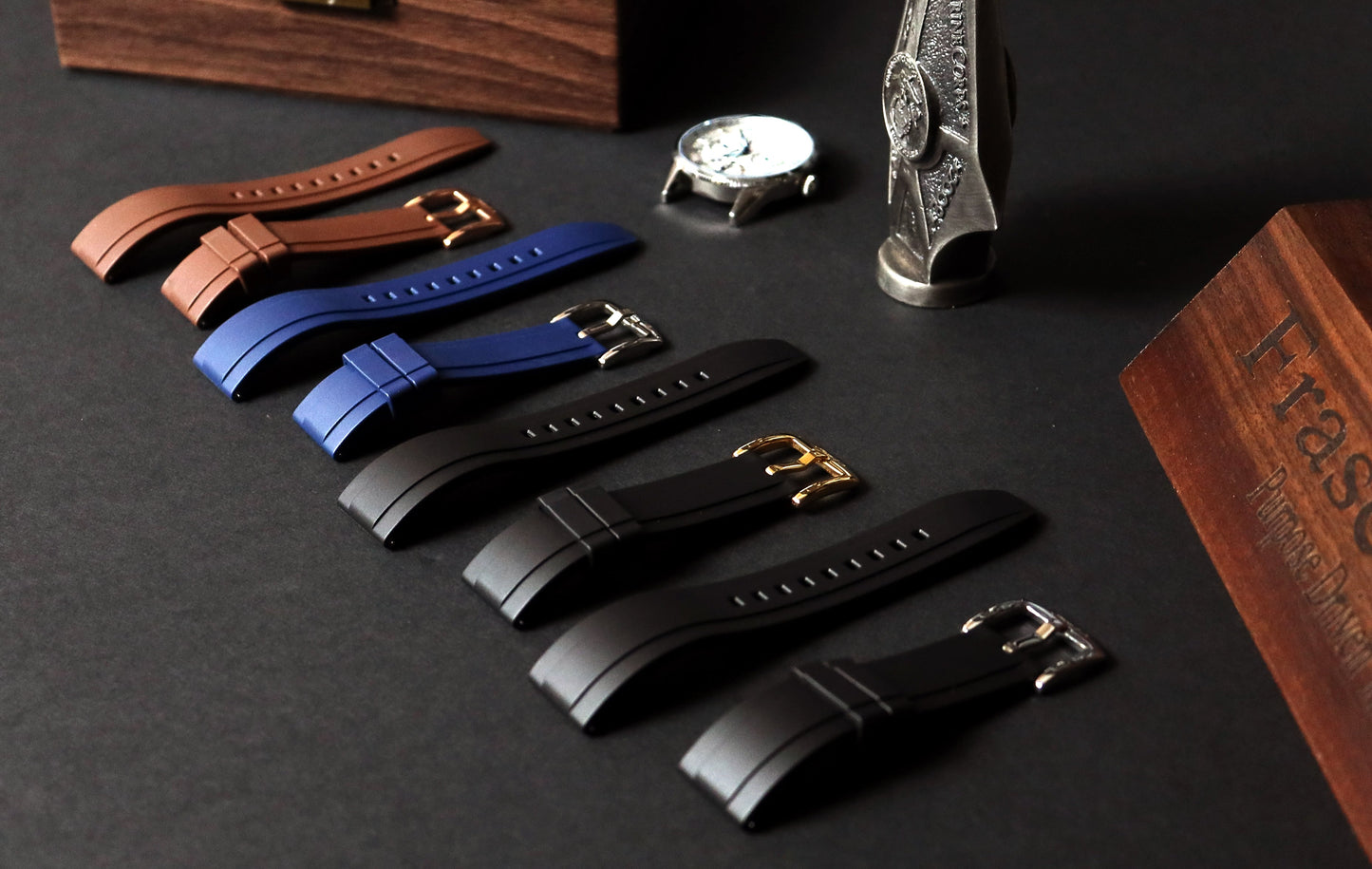 Set of watch straps in brown, blue, and black on a dark surface with a watch and box in the background.