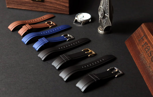 Set of watch straps in brown, blue, and black on a dark surface with a watch and box in the background.