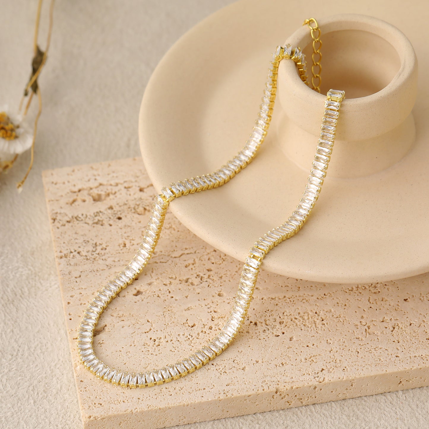 Gold and crystal necklace draped over a beige ceramic dish on a textured beige surface