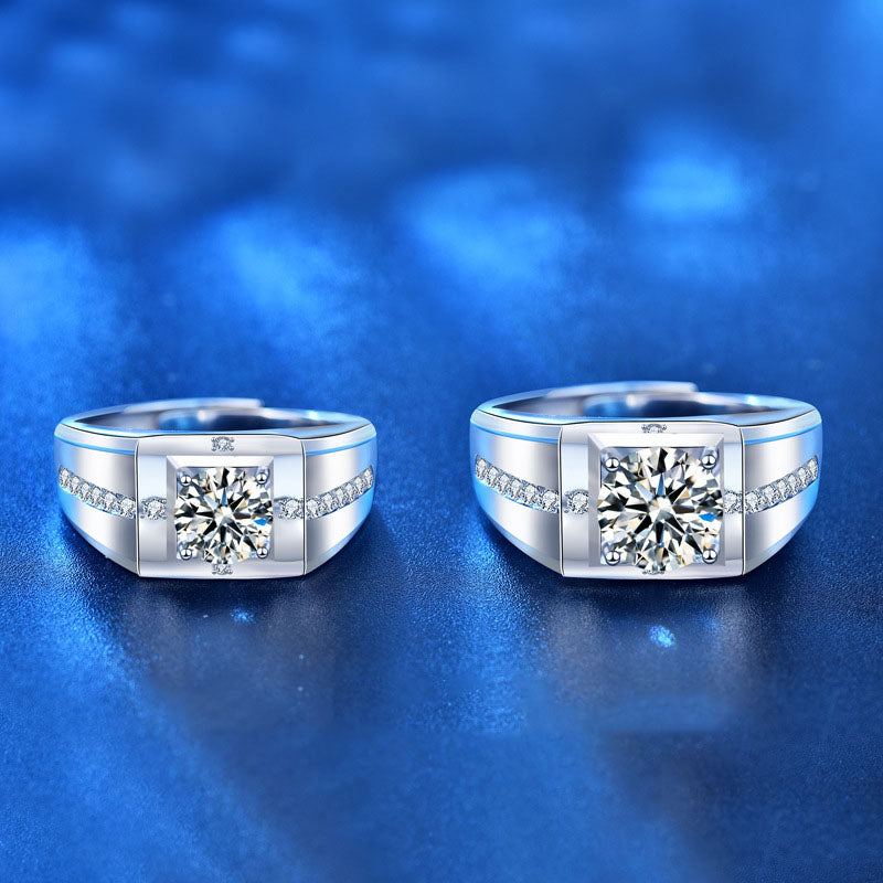 Two silver-toned men's ring bands with square-cut clear center stones and small side accent stones, displayed on a glossy blue background.