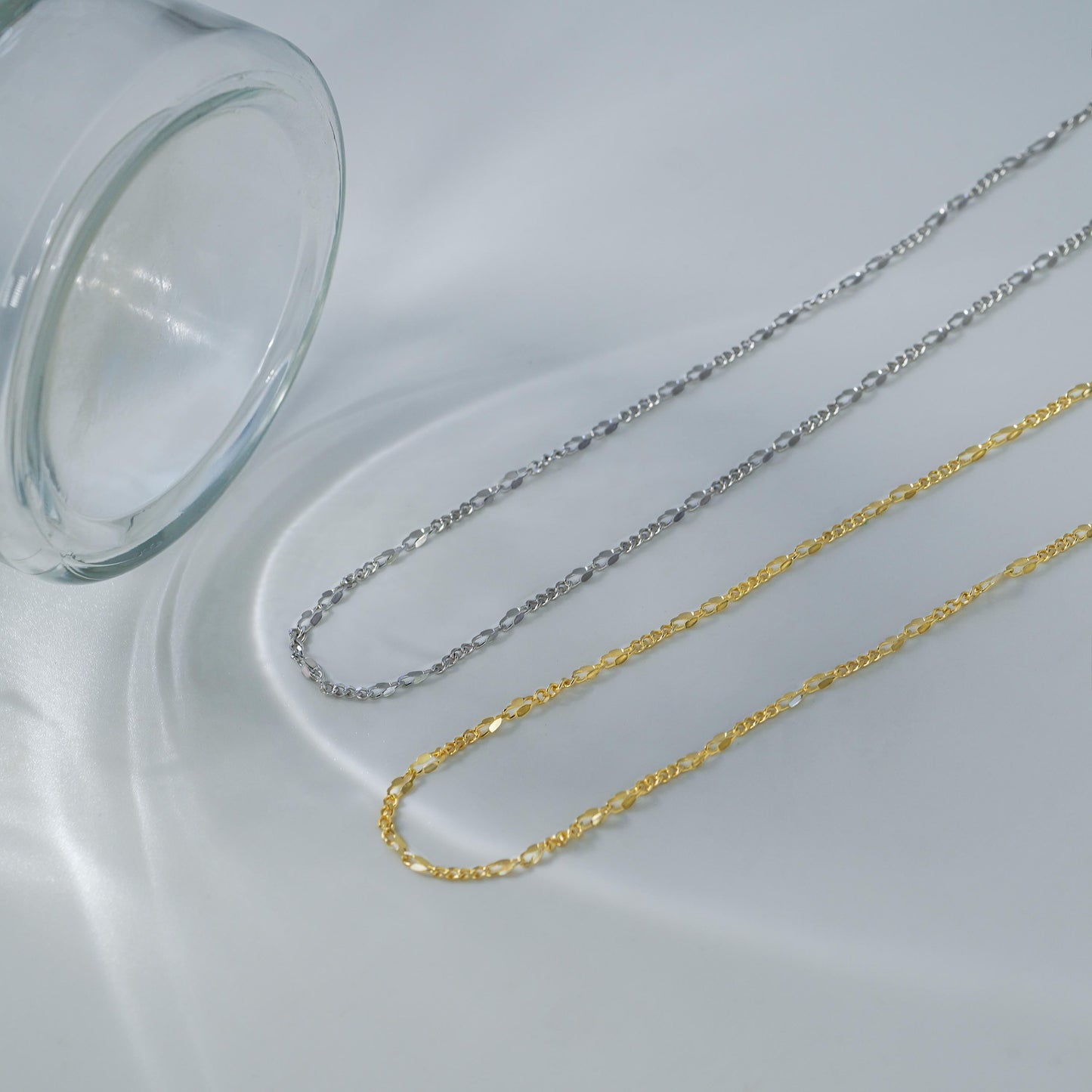Two gold and silver chains on a white surface with a clear glass container.