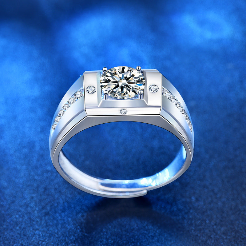 A silver-toned men's ring featuring a round-cut center stone and small accent stones along the shoulders, set against a textured blue background.