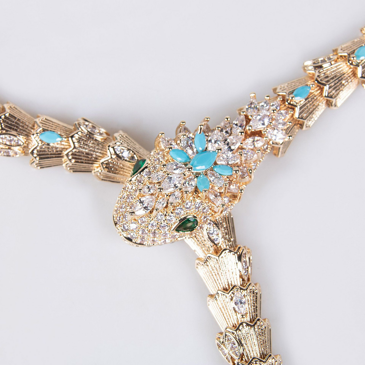 Close-up of a gold snake necklace head with turquoise blue and clear stones on a white background.