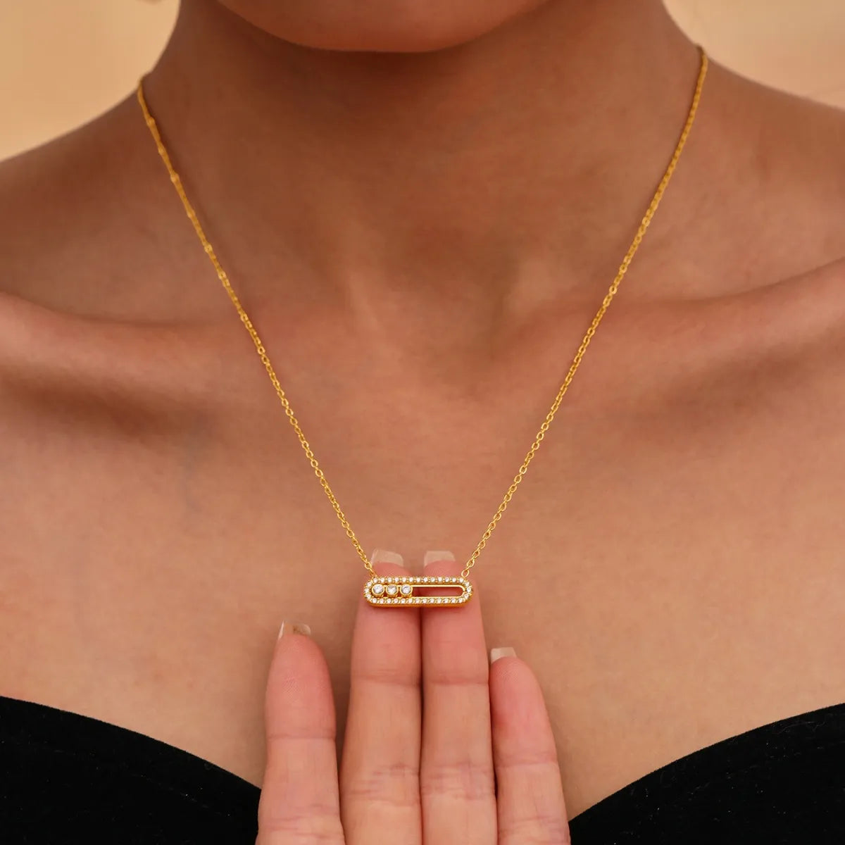 Gold necklace with a bar pendant held by a hand against a beige background