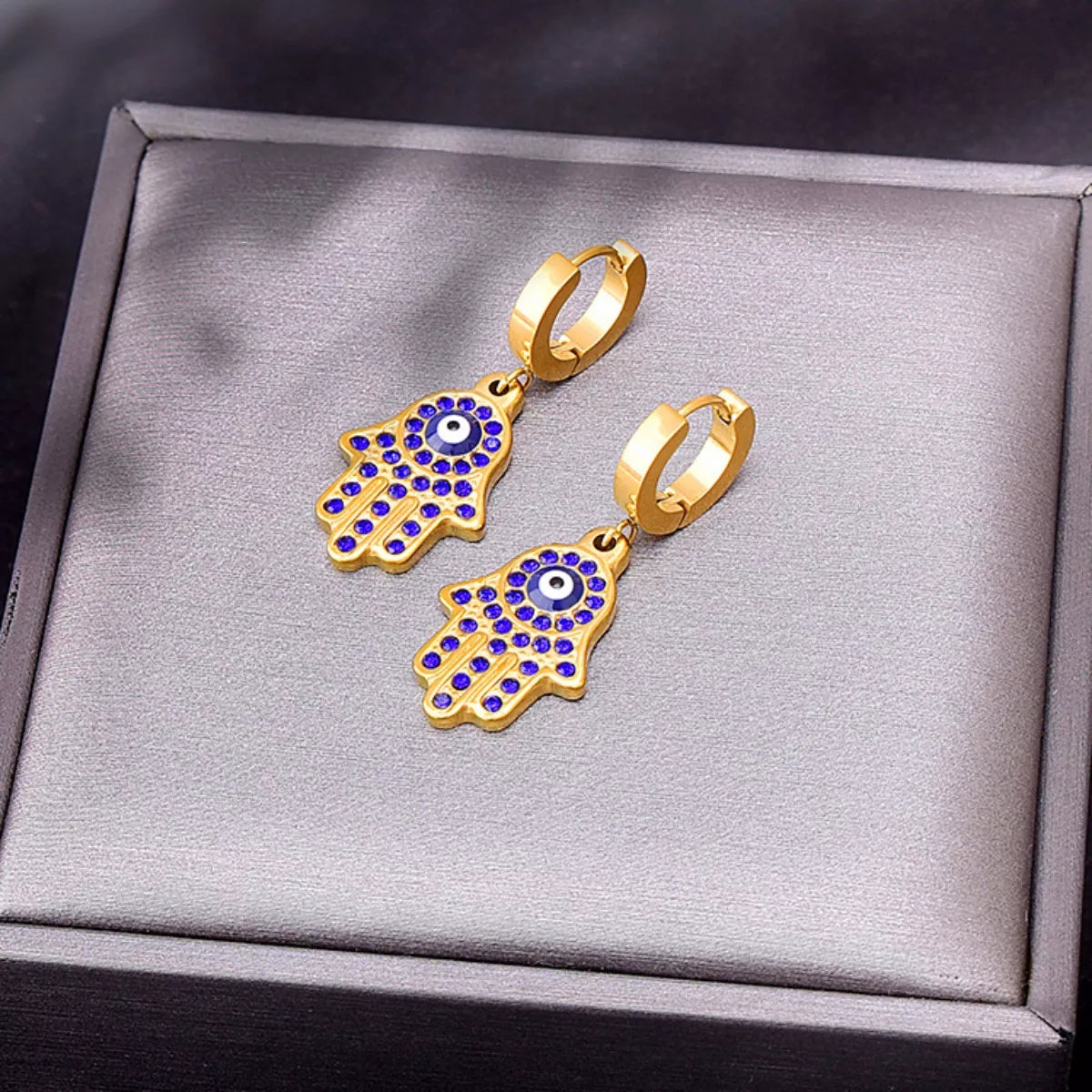 Gold hamsa hand earrings with blue accents on a gray jewelry box