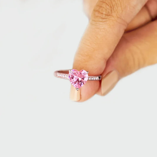 Heart-shaped pink gemstone ring held between fingers on a light background