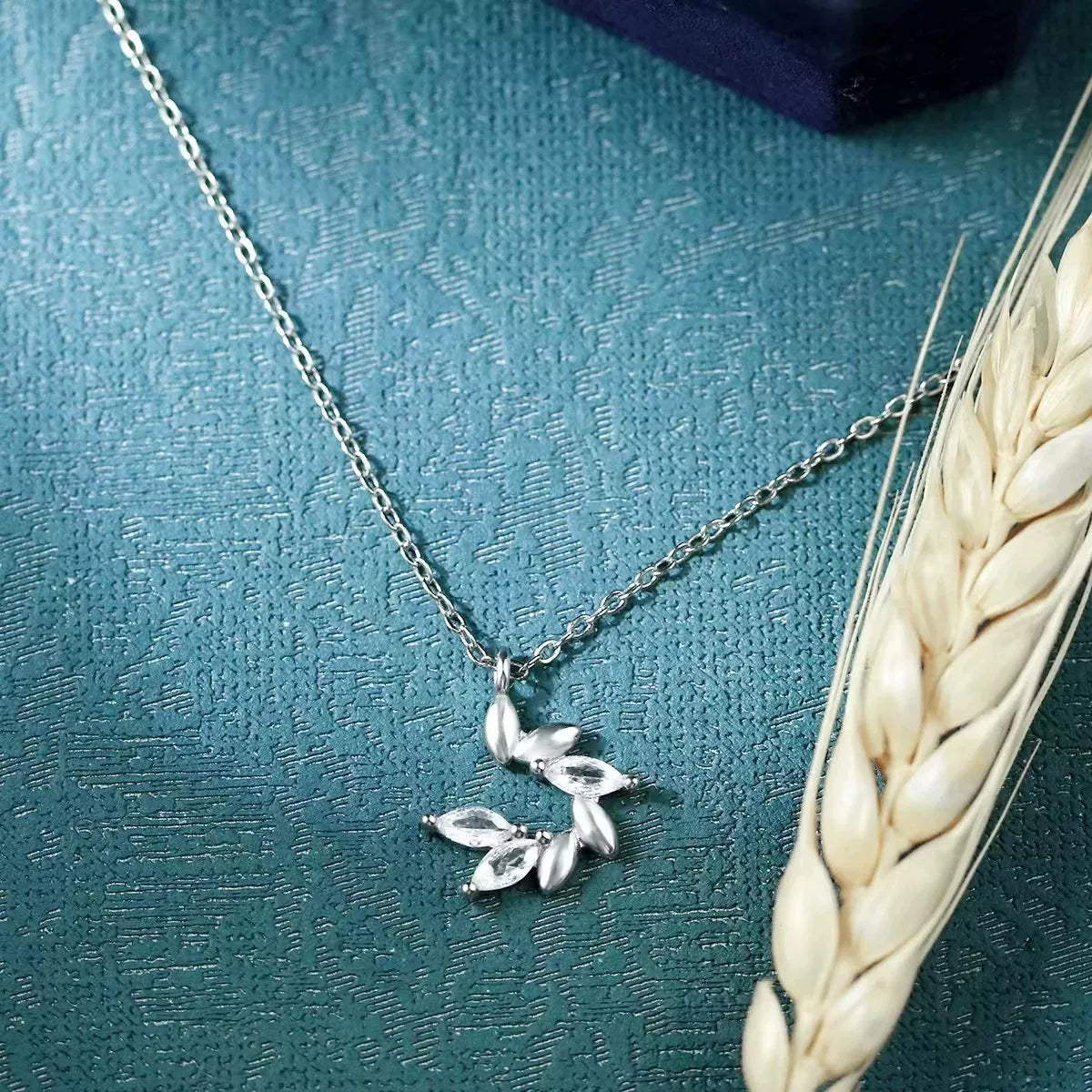 Silver necklace with leaf pendant on a textured teal surface with wheat stalk.