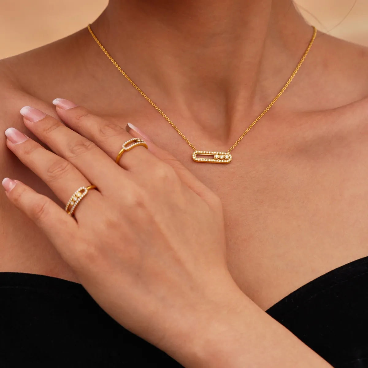 Gold necklace and rings on a person's hand and chest against a neutral background
